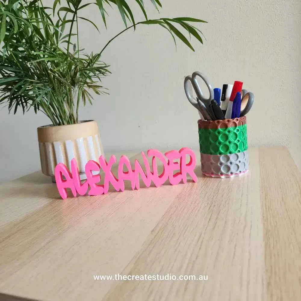 Custom name decor gift in pink spelling 'ALEXANDER' on a wooden table with office supplies