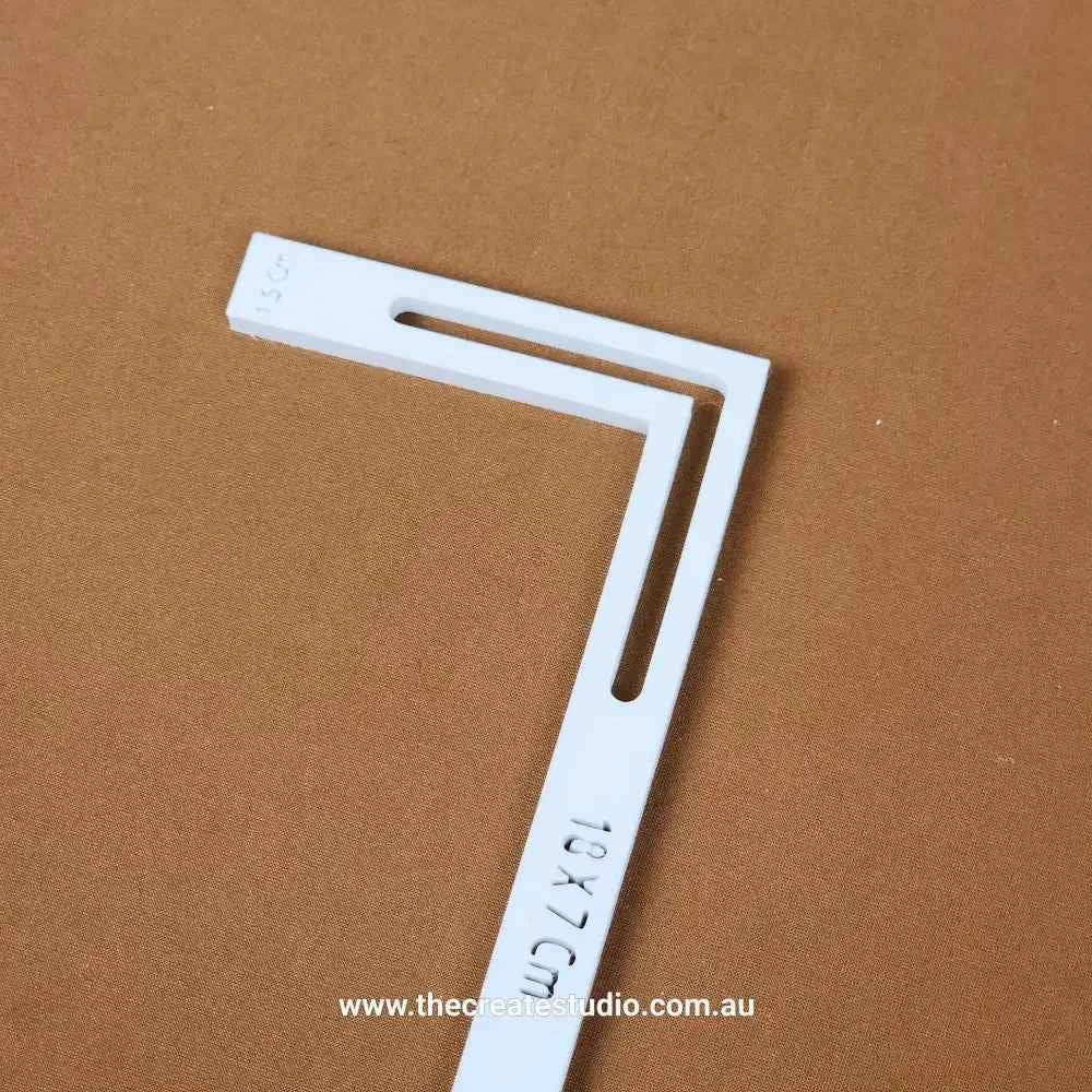 White plastic L-piece bookbinding tool with elongated slots and printed measurements on brown background