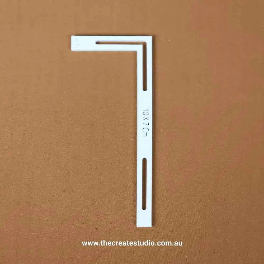 White plastic L-piece bookbinding tool with elongated slots and 10x7cm marking on brown background